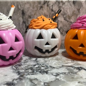 Puede incluir: Tres recipientes con forma de calabaza de Halloween con coberturas de crema batida y pajitas. Las calabazas son rosas, blancas y naranjas, cada una con una cara de Jack-o'-lantern tallada. Acentos negros y un asa completan el diseño.