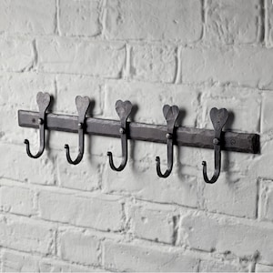 May include: A dark metal coat rack with five hooks, mounted on a white brick wall. The hooks have a heart-shaped design and are attached to a textured metal bar. The rack is designed for wall mounting.