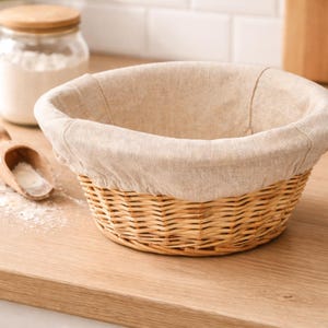 May include: A round wicker bread proving basket with a beige linen lining. The basket is on a light wood surface, with a wooden spoon and a jar of flour in the background. The basket is empty.