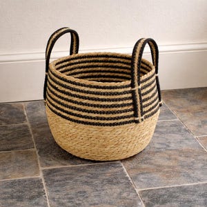 May include: Round woven basket with black horizontal stripes and two black handles. The basket is made of natural-coloured jute material and is sitting on a tiled floor. The basket is designed for storage.