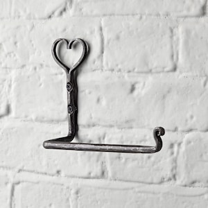May include: A wrought iron toilet paper holder with a heart-shaped top. The holder is mounted on a white brick wall. The metal has a dark, rustic finish, and the design includes a curved end for the paper roll.