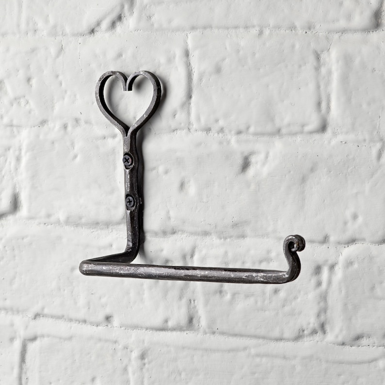 May include: A wrought iron toilet paper holder with a heart-shaped top. The holder is mounted on a white brick wall. The metal has a dark, rustic finish, and the design includes a curved end for the paper roll.
