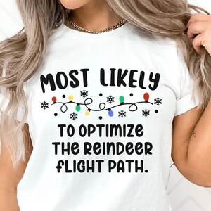 May include: White t-shirt with black text that reads "Most Likely To Optimize The Reindeer Flight Path." The text is surrounded by a string of colorful Christmas lights and snowflakes.