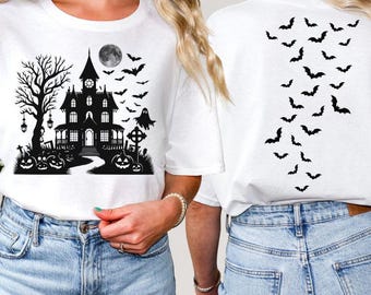 May include: White t-shirt featuring a black Halloween haunted house design with a full moon, bats, and pumpkins. The back of the shirt has a pattern of black bats. A spooky and festive design for Halloween.