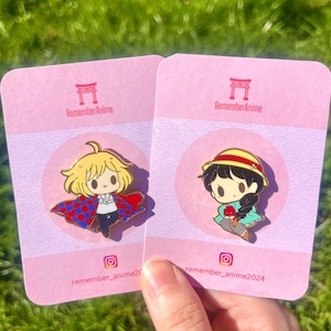 Könnte beinhalten: Zwei Emaille-Anstecknadeln auf rosa Karten. Eine Anstecknadel zeigt einen blonden Charakter mit rotem und blauem Umhang. Die andere Anstecknadel zeigt einen Charakter mit gelbem Hut und schwarzen Haaren. Die Karten haben den Text "RememberAnime" und einen Instagram-Handle.