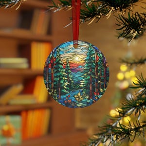 May include: A round Christmas ornament with a stained glass design of a forest scene. The ornament features a sunset with a bright orange sky, blue mountains, and two tall green trees.