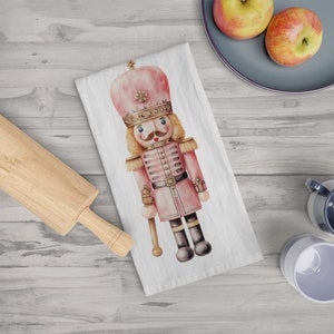 May include: White kitchen towel featuring a watercolor illustration of a pink nutcracker soldier. The nutcracker has a gold crown, a mustache, and is holding a staff. The towel is on a light wood surface, with a rolling pin, apples, and mugs.