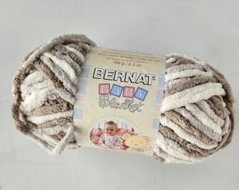 May include: A skein of Bernat Baby Blanket yarn featuring a soft, chenille-style texture in alternating stripes of cream and light brown. The central paper label displays the brand name, weight of 100 g / 3.5 oz, and a graphic of a baby.