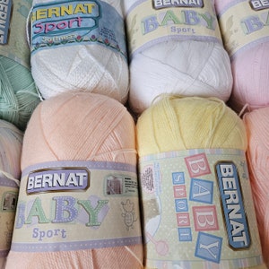 K&ouml;nnte beinhalten: F&uuml;nf Kn&auml;uel Bernat Baby Sport Garn in verschiedenen Pastellfarben, darunter Rosa, Pfirsich, Gelb, Wei&szlig; und Gr&uuml;n. Jedes Kn&auml;uel hat ein Etikett mit dem Bernat Baby Sport Logo und den Worten "Weichheit" und "Kostenloses Muster auf dem Etikett".