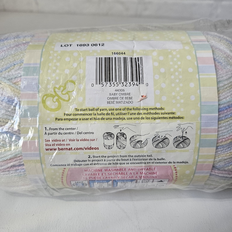 K&ouml;nnte beinhalten: Nahaufnahme eines Garnetiketts mit dem Text "LOT 1693 0612" und "BABY OMBRE" in einem hellblauen und wei&szlig;en Farbschema. Das Etikett enth&auml;lt auch Anweisungen zum Starten einer Garnkugel und Waschanweisungen.