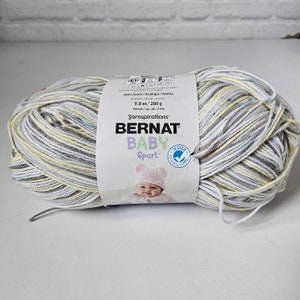 K&ouml;nnte beinhalten: Eine Kn&auml;uel Bernat Baby Sport Garn in einem mehrfarbigen Muster aus Wei&szlig;, Grau, Gelb und Blau. Das Etikett lautet "100% Acrylic/Acrylique/Acrilico", "9.8 oz/280 g", "1004 yds/vgs/yds/915 m", "Yarnspirations", "BERNAT", "BABY", "Sport", und "TESTED".