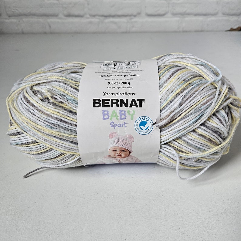 K&ouml;nnte beinhalten: Eine Kn&auml;uel Bernat Baby Sport Garn in einem mehrfarbigen Muster aus Wei&szlig;, Grau, Gelb und Blau. Das Etikett lautet "100% Acrylic/Acrylique/Acrilico", "9.8 oz/280 g", "1004 yds/vgs/yds/915 m", "Yarnspirations", "BERNAT", "BABY", "Sport", und "TESTED".