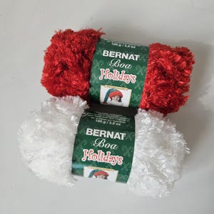 May include: Two skeins of Bernat Boa Holidays yarn, one red and one white. Each skein is labeled with "100 g / 3.5 oz". The yarn is fluffy and soft, perfect for holiday crafting projects.