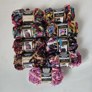 May include: Nine skeins of Bernat Boa yarn arranged in a grid. Each skein features a textured, multi-colored fringe design with a central paper label displaying the brand name, product line, and a photo of a person wearing a garment made from the yarn.