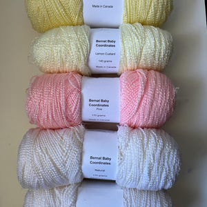 Könnte beinhalten: Fünf Knäuel Bernat Baby Coordinates Garn, vertikal gestapelt. Die Etiketten zeigen die Farben: Gelb (140g), Zitronencreme (140g), Rosa (170g), Natur (170g) und Weiß. Jedes Knäuel besteht aus einem strukturierten Garnstrang.