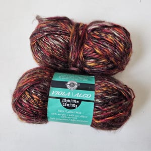 May include: Two skeins of variegated yarn in shades of deep red, orange, black, and white. A teal paper label wraps around the center, displaying the text Loops & Threads, Viola | Alto, 213 yds, 3.5 oz, 64% acrylic, and 36% polyester.
