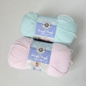 May include: Two skeins of Loops & Threads Snuggly Wuggly baby sport yarn. One is a light blue, the other is pink. The yarn is soft and fluffy, perfect for baby projects. The labels feature a logo and the words "Snuggly Wuggly baby sport".