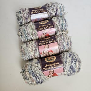 May include: Four skeins of Homespun Thick & Quick yarn stacked vertically. Each skein features a textured, multi-colored blend of cream, blue, and purple fibers. The paper labels display the brand name and a photograph of a person holding yarn.