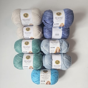 May include: Assortment of Lion Brand Rewind Tape Yarn in various colors, including cream, blue, teal, and light blue. Each skein is labeled with the "REWIND TAPE YARN" logo and product information. Ideal for knitting and crochet projects.