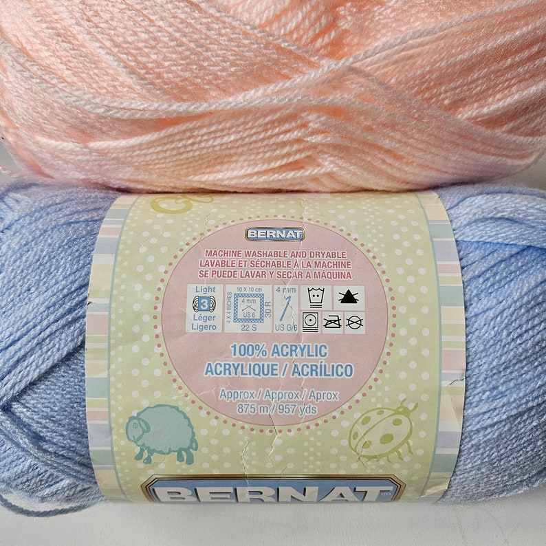 K&ouml;nnte beinhalten: Zwei Kn&auml;uel Bernat-Garn, eines hellrosa und eines hellblau. Das Garn ist 100% Acryl und maschinenwaschbar und trocknergeeignet. Das Etikett enth&auml;lt auch das Garngewicht, die L&auml;nge und die Pflegehinweise.