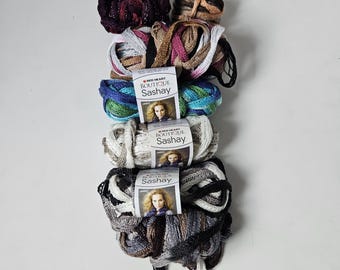 May include: A vertical stack of five skeins of Red Heart Boutique Sashay yarn. The colors include deep burgundy, tan, blue and green, white and grey, and a multi-colored mix of black, white, and grey. Each skein features a paper label with text.