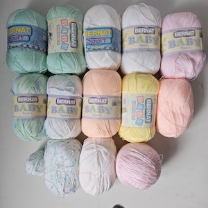 K&ouml;nnte beinhalten: Eine Sammlung von 11 Garnkn&auml;uel in verschiedenen Pastellfarben, darunter Rosa, Blau, Gr&uuml;n, Gelb und Wei&szlig;. Jedes Kn&auml;uel tr&auml;gt das Etikett "Bernat Baby Sport".