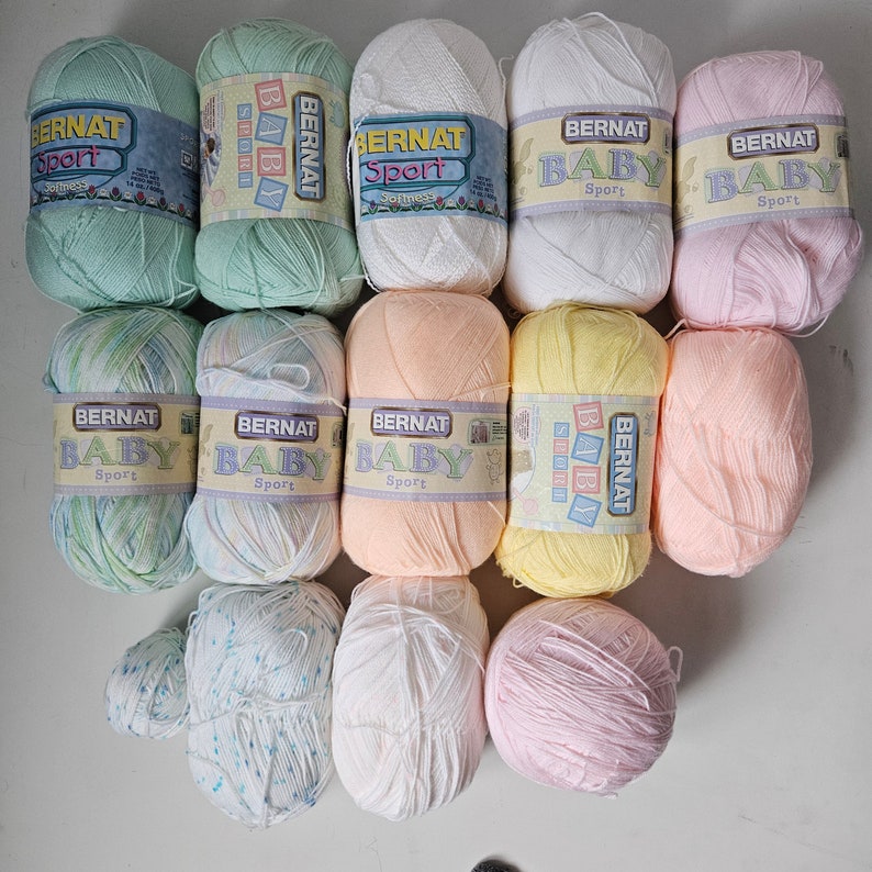 K&ouml;nnte beinhalten: Eine Sammlung von 11 Garnkn&auml;uel in verschiedenen Pastellfarben, darunter Rosa, Blau, Gr&uuml;n, Gelb und Wei&szlig;. Jedes Kn&auml;uel tr&auml;gt das Etikett "Bernat Baby Sport".