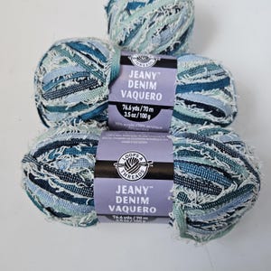 May include: Three balls of Jeany Denim Vaquero yarn by Loops & Threads in shades of blue, teal, and white. Each ball features a textured, frayed appearance and a paper label stating 76.6 yds, 70 m, 3.5 oz, 100 g, and 70% acrylic composition.