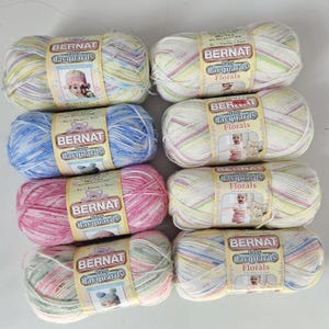 May include: Eight skeins of Bernat Baby Jacquards Florals yarn in various colors. The yarn is a mix of pastel shades, including pink, blue, yellow, and green. Each skein is wrapped with a label featuring a baby and a teddy bear.