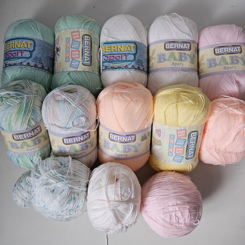 K&ouml;nnte beinhalten: Eine Sammlung von zehn Kn&auml;uel Bernat Baby Sport Garn in verschiedenen Pastellfarben, darunter Pfirsich, Gelb, Rosa, Wei&szlig; und Gr&uuml;n. Das Garn ist mit dem Markennamen und den Worten "Baby Sport" gekennzeichnet.