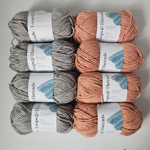 May include: Eight skeins of Loops & Threads Ringspun acrylic blend yarn in two colours: grey and peach. Each skein has a label with the brand name and "Filée à l'Anneau" printed on it. The yarn is arranged in two columns.