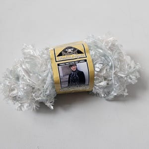 May include: A skein of white and light blue eyelash yarn with a label that reads "Phentex Fashion Eight". The label features a photo of a woman wearing a scarf. The yarn is fluffy and textured, suitable for knitting or crochet projects.