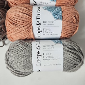 Peut inclure: Gros plan sur plusieurs pelotes de fil Ringspun, mélange acrylique, en tons pêche et gris. Chaque pelote porte une étiquette avec le nom de la marque, le type de fil et le texte "Filée à l'Anneau". Les étiquettes indiquent également 149 m / 170 g.