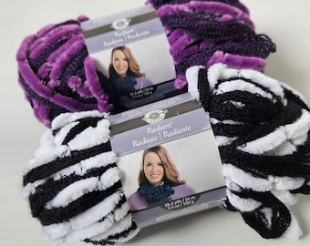 May include: Three skeins of Radiant yarn in purple, black and white. The purple yarn has a fluffy texture with small pom-poms. The black and white yarn is striped. Each skein has a label with the text "Radiant" and "26.4 yds / 24 m".