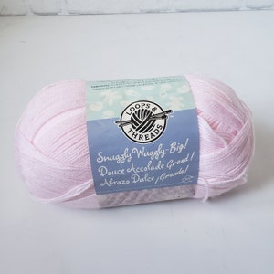 May include: A large skein of soft pink yarn with a label that reads "Loops & Threads Snuggly Wuggly-Big!" in multiple languages. The yarn is a light pastel color, ideal for baby blankets and other craft projects.