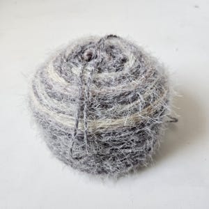 May include: A round ball of fuzzy yarn featuring a variegated color palette of charcoal gray, light gray, and cream. The texture is highly fibrous and hairy, with loose strands winding around the spherical shape, creating a soft, multi-tonal appearance.