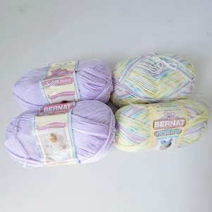 May include: Four skeins of baby yarn in pastel colors. Two skeins are solid lavender, labeled "Softer Baby". The other two are variegated, with yellow, blue, and lavender, labeled "Bernat Baby Jacquards". Ideal for baby blankets and other projects.