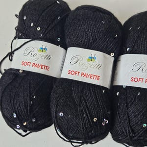 May include: Close-up of three black yarn skeins with embedded sequins. Each skein has a white label with the text "Rozetti SOFT PAYETTE". The yarn is tightly wound, and the sequins add a touch of sparkle to the dark material.