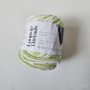 May include: A ball of Loops & Threads Everyday Cotton yarn featuring a variegated pattern of white and light green strands. The white paper label displays the brand name, the text Everyday Cotton, Coton Quotidien, and 86 M / 56.7 G.