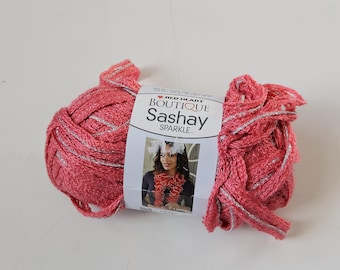May include: A skein of Red Heart Boutique Sashay Sparkle yarn in a vibrant coral pink color with metallic silver threading. The center label features the product name and a photograph of a person wearing a ruffled scarf made from this yarn.