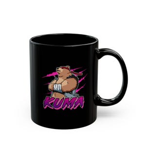 Puede incluir: Taza de café de cerámica negra con un gráfico de un oso marrón con una cinta roja en la cabeza y una camisa azul. El texto "KUMA" está impreso en rosa debajo del oso.