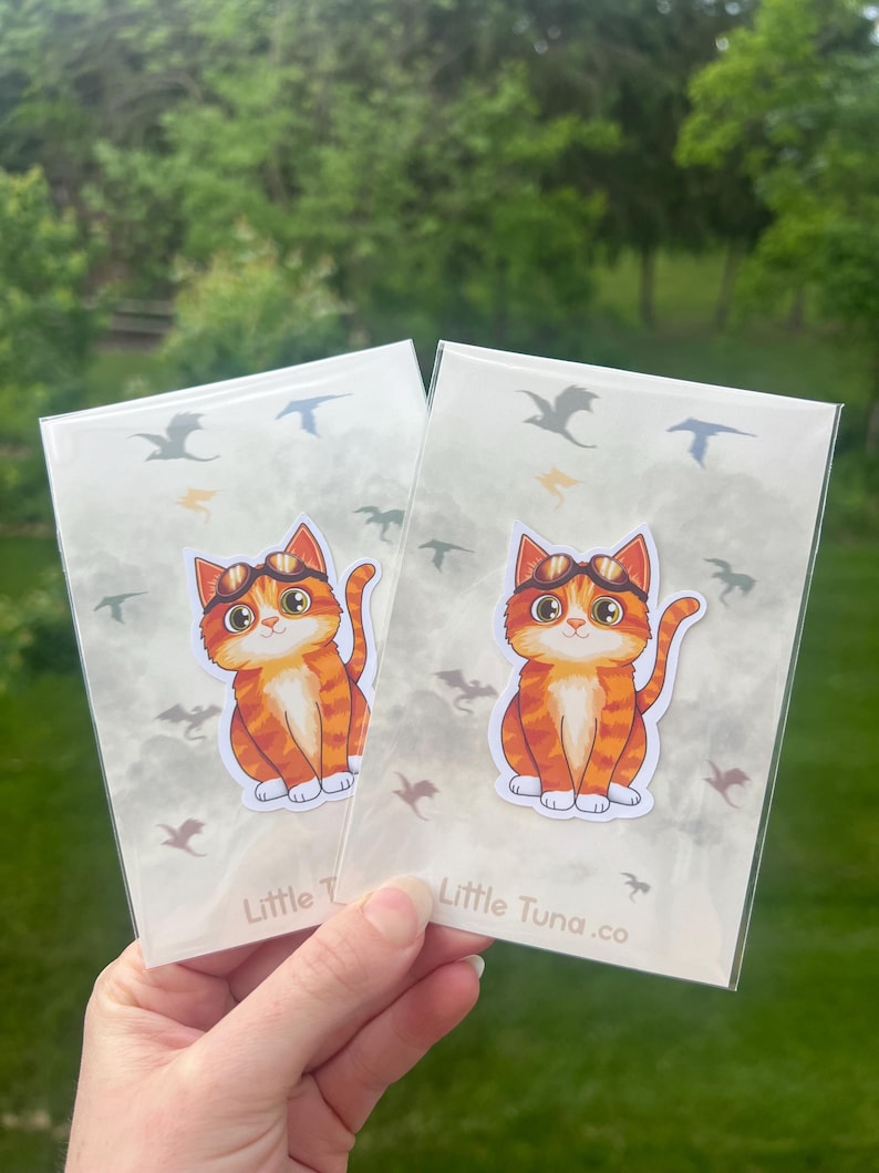 May include: Two stickers featuring an orange tabby cat wearing goggles. The cat is sitting upright with its tail curled. The stickers are in clear plastic sleeves with a cloudy background and bird and dragon illustrations. The text "Little Tuna.co" is visible.