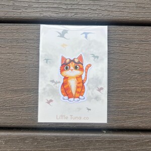 May include: A sticker featuring a cartoon orange tabby cat wearing goggles, surrounded by flying dragon silhouettes. The sticker has the text "Little Tuna.co" at the bottom, and is on a white background.
