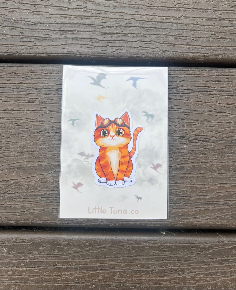 May include: A sticker featuring a cartoon orange tabby cat wearing goggles, surrounded by flying dragon silhouettes. The sticker has the text "Little Tuna.co" at the bottom, and is on a white background.