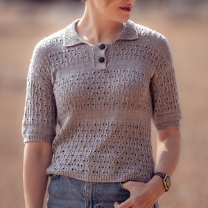 May include: A woman wearing a light gray knitted sweater with a collared neckline and short sleeves. The sweater has a delicate openwork pattern. She is also wearing blue jeans and a black watch.