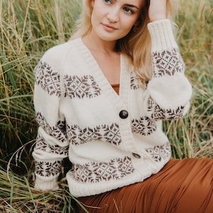 May include: A woman wearing a white and brown patterned cardigan with a brown skirt. She is sitting in a field of tall grass.