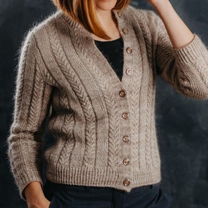 Op de afbeelding: Een beige kabelgebreid vest met een V-hals en knoopsluiting. De trui met lange mouwen heeft een textuurontwerp en wordt gecombineerd met een donkerblauwe broek. Het vest heeft een neutrale kleur en lijkt gemaakt van een zacht, warm materiaal.