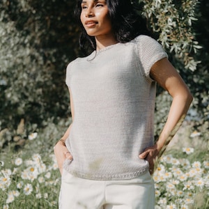 Puede incluir: Una mujer con un top de punto gris claro de manga corta y pantalones blancos se encuentra en un campo de margaritas blancas.