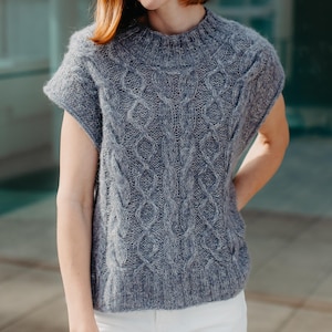 May include: A slate blue sleeveless knit sweater featuring a prominent vertical cable knit pattern, a ribbed crew neckline, and a ribbed hem. The garment is constructed with a textured, slightly fuzzy yarn and has a relaxed, boxy silhouette.