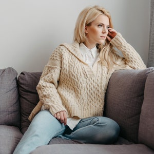 May include: A woman wearing a cream-colored cable knit sweater with a white collared shirt underneath, sitting on a gray couch. She is looking off to the side and has her hand on her cheek.
